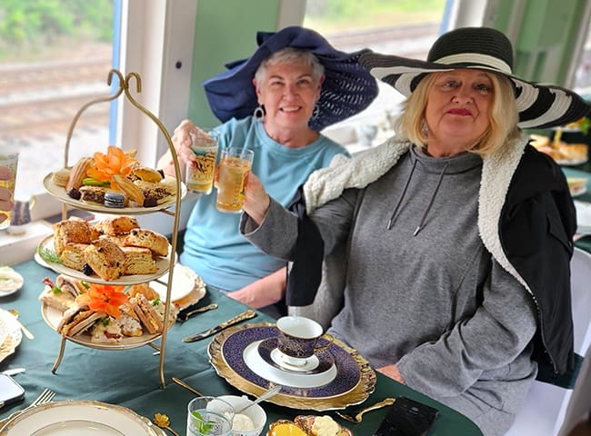 Two elderly women in wide-brim hats raise drinks at a table set with a three-tier pastry and sandwich stand for afternoon tea.