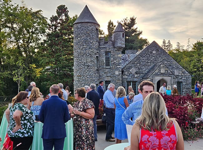 Guests mingle outside a stone castle-style building with towers during a daytime formal event.