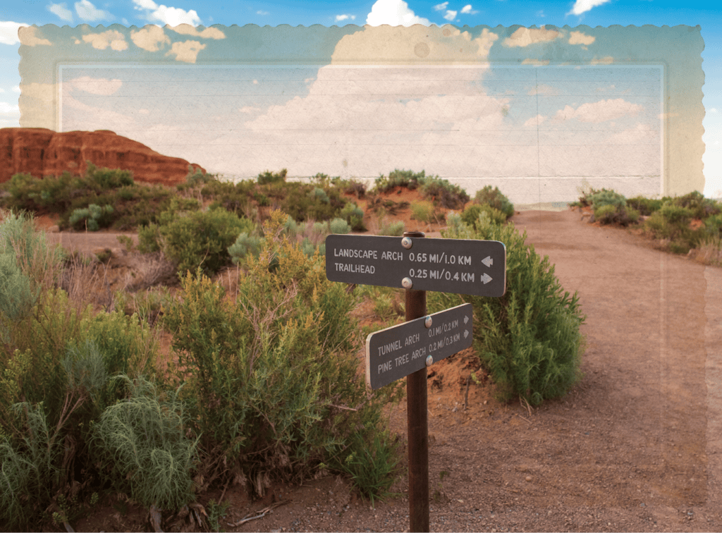 Desert trail signpost listing distances to Landscape Arch and Trailhead, with scrubby bushes and red rock formations in the distance under a blue sky.
