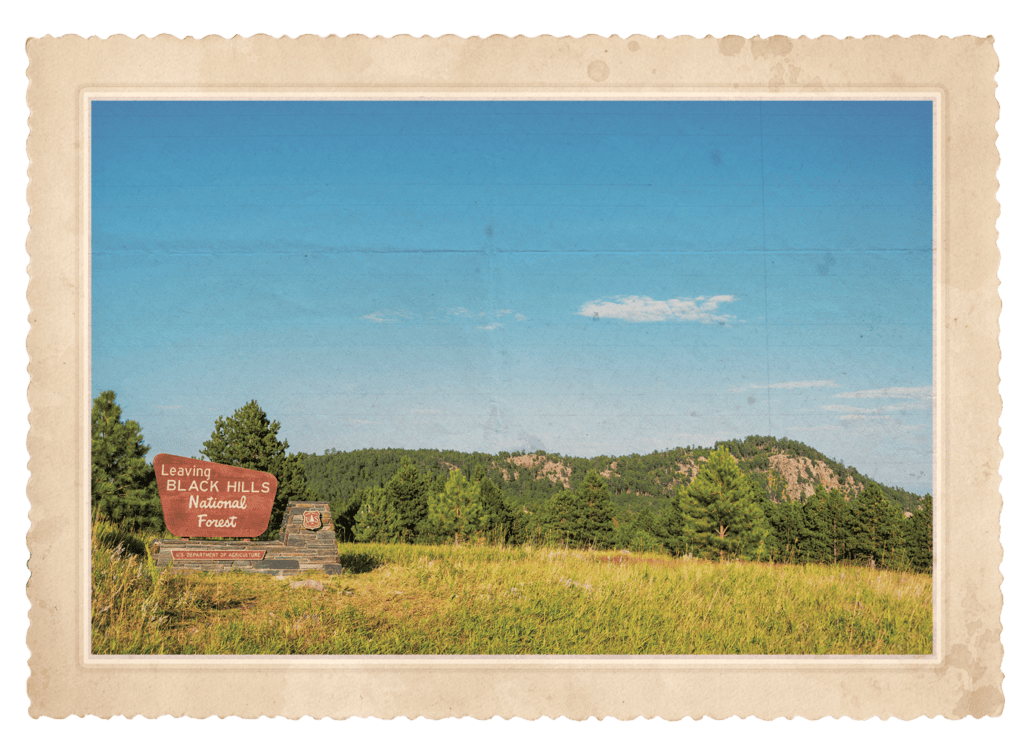 Sign reading 'Leaving Black Hills National Forest' beside a grassy field with pine trees and distant hills under a blue sky, in a vintage photo frame.