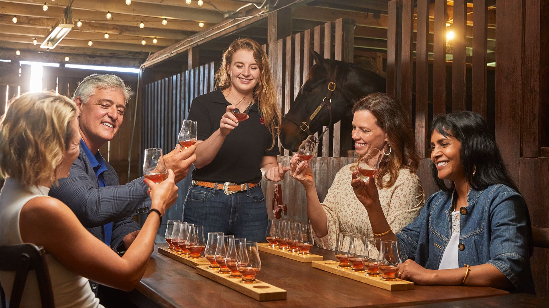 Group of five adults toasting with whiskey glasses around a tasting table in a rustic barn setting with horse stalls in the background.