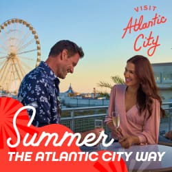 Two people chatting with drinks on a boardwalk near a large Ferris wheel, with overlay text: 'Summer' and 'The Atlantic City Way' and a 'Visit Atlantic City' logo in the corner.