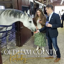 Two adults feed a white horse in a barn stall; Oldham County Tourism & Conventions branding visible.