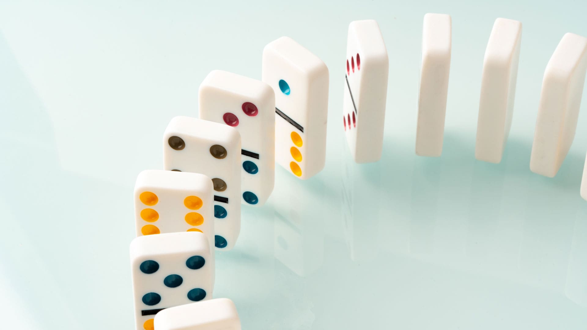 Row of white domino tiles lined up and tipping over in a chain reaction on a light blue surface to illustrate domino effect